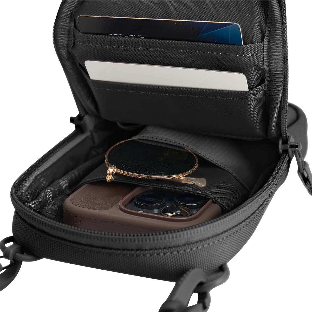 Borsa nera aperta con carte di credito, una carta bianca, occhiali da sole e un telefono in una custodia marrone.