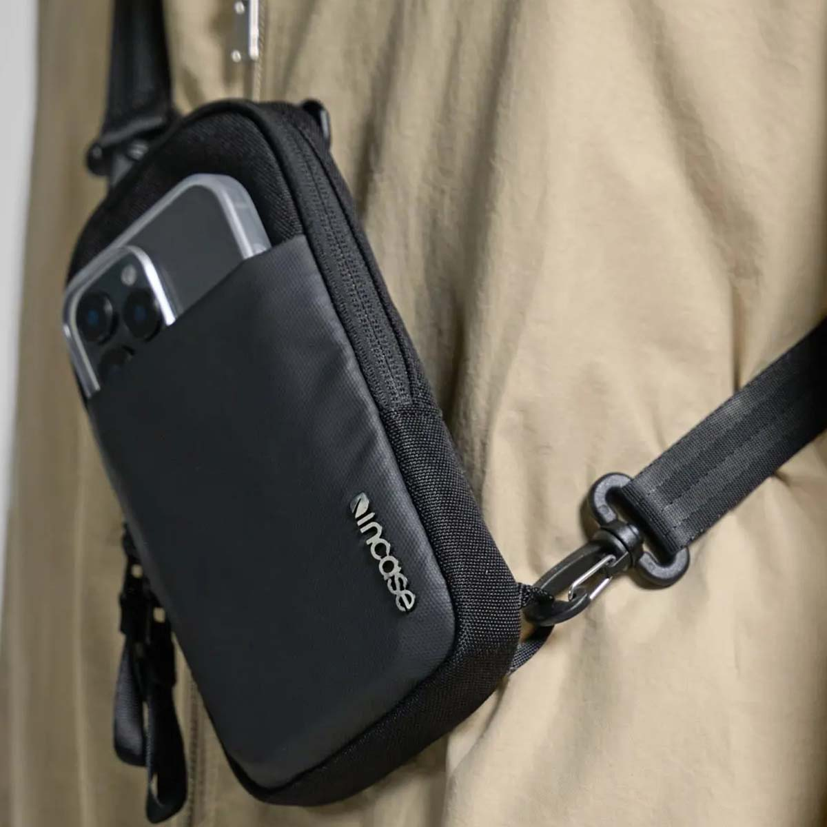 Una borsa nera a tracolla con un telefono in tasca. La borsa è appesa a una persona con una giacca beige.