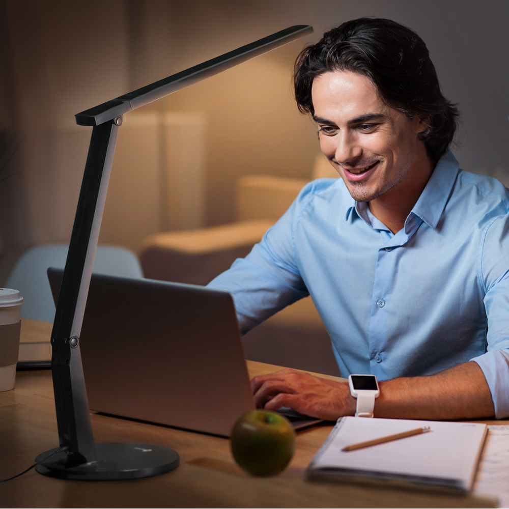 Ein Mann lächelt, während er an einem Laptop an einem Schreibtisch arbeitet. Eine schwarze Lampe scheint auf den Schreibtisch.