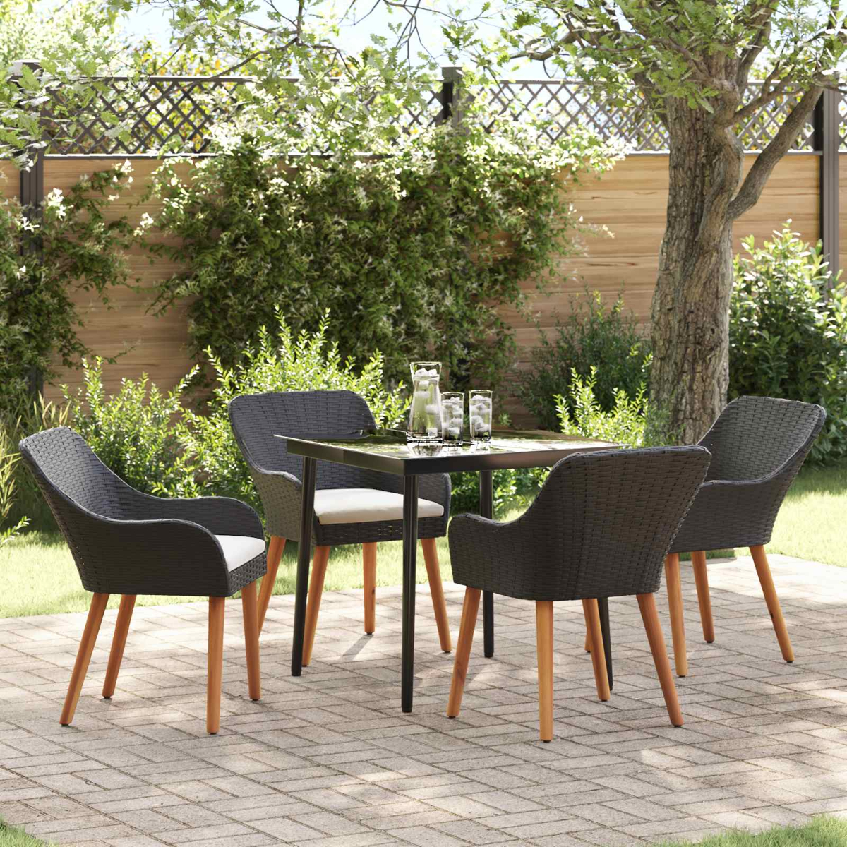 Juego de patio al aire libre con cuatro sillas tejidas de color gris oscuro, una mesa cuadrada con vasos y una jarra. El exuberante follaje verde rodea.