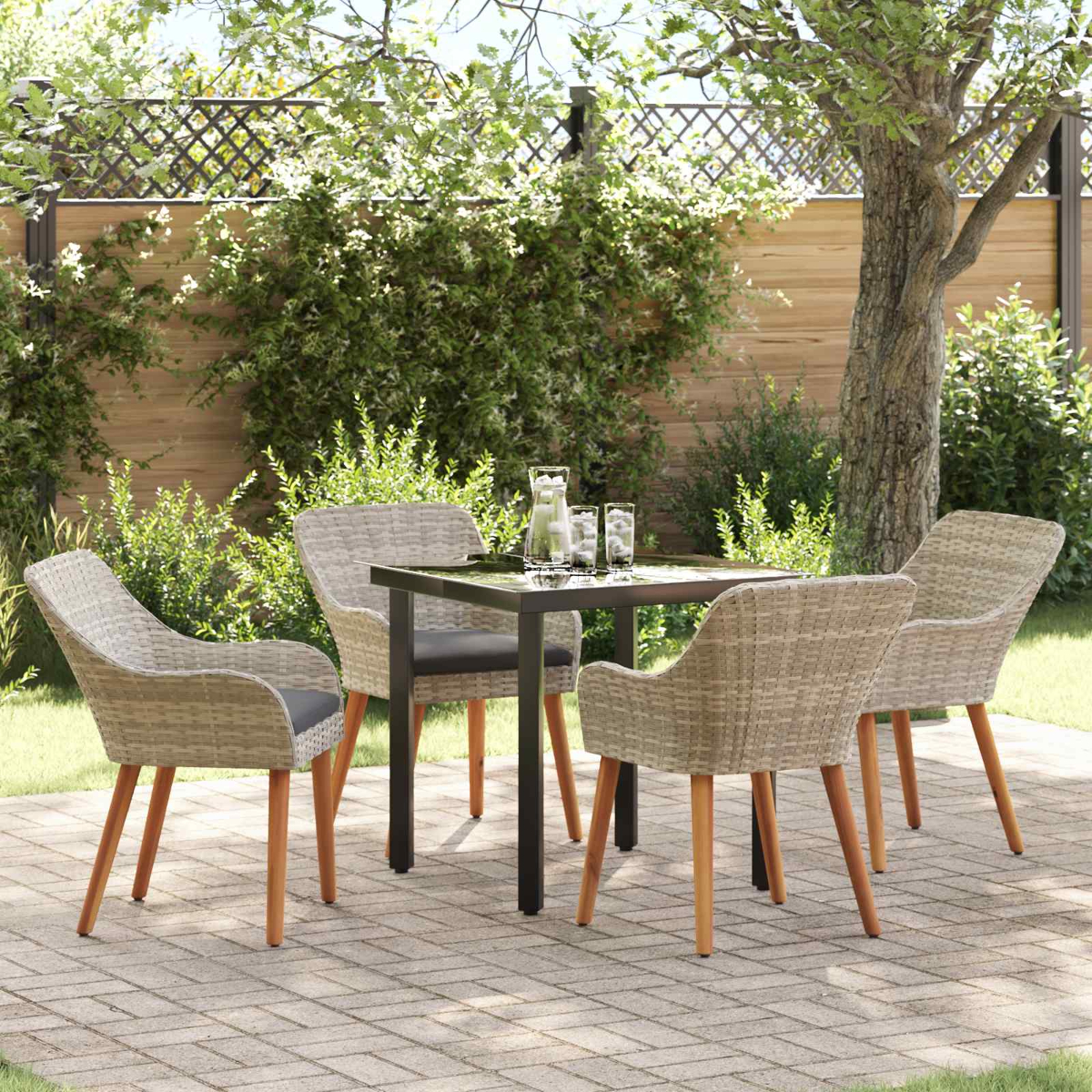Terrassengarnitur mit vier Korbstühlen, quadratischem Tisch und Getränken. Grünes Laub und ein Baum im Hintergrund. Gepflasterter Boden.