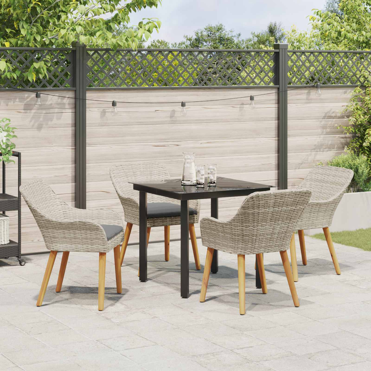 Terrassengarnitur im Freien. Quadratischer schwarzer Tisch mit vier Korbstühlen. Holzzaun und Grün im Hintergrund. Lichterkette darüber.