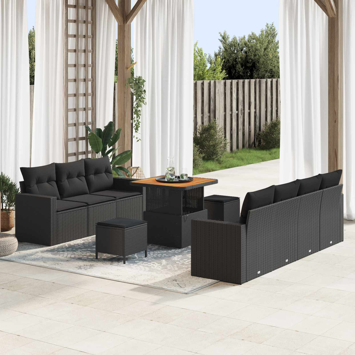 Terrassenmöbel-Set mit schwarzen Korbs Sofas, Hocker, Tisch und weißen Vorhängen. Ein Holzzaun und grüner Rasen im Hintergrund.