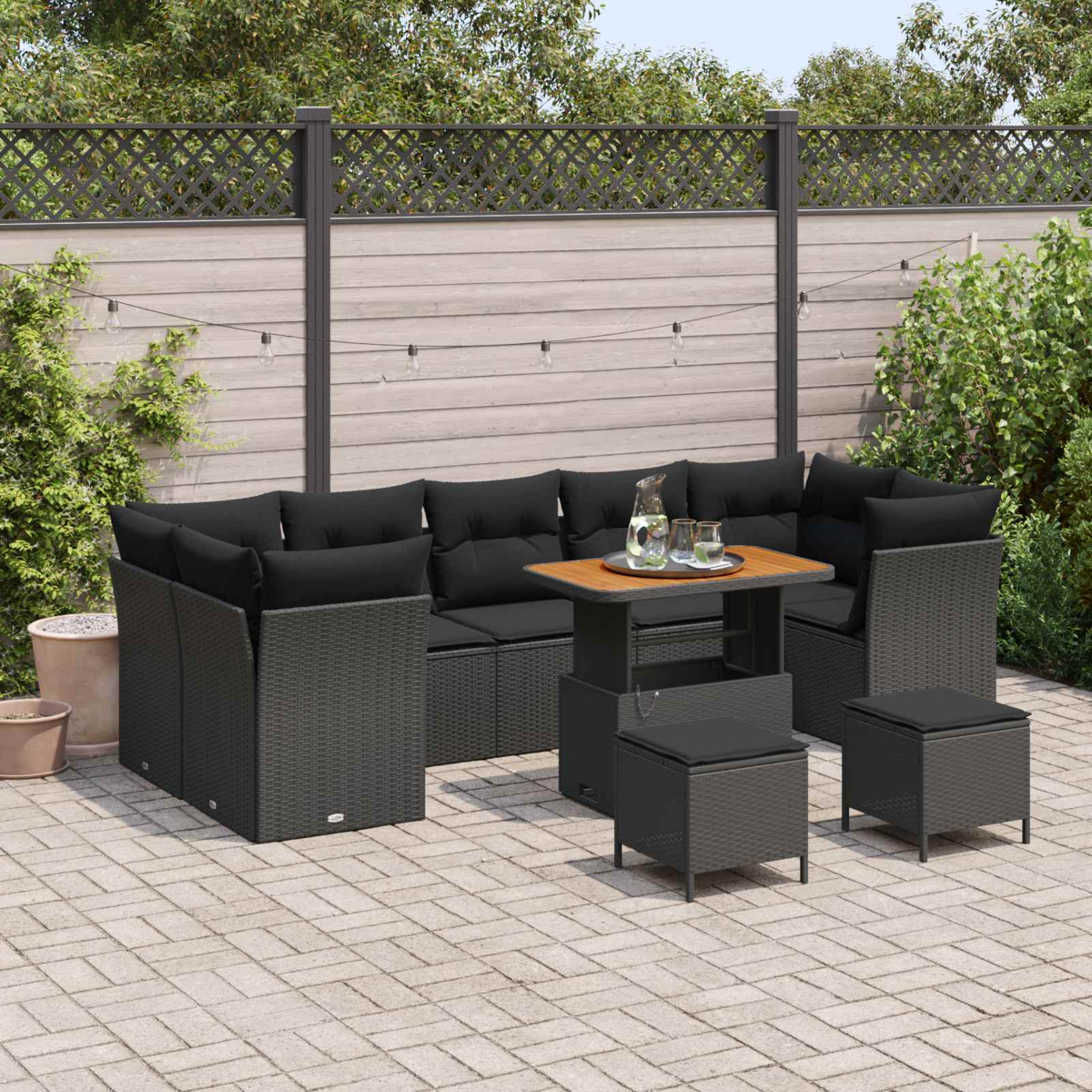 Garten-Terrassenset mit schwarzen Kissen, Tisch mit Gläsern und Krug, graue Stühle auf einer Backsteinterrasse, vor einem Holzzaun.
