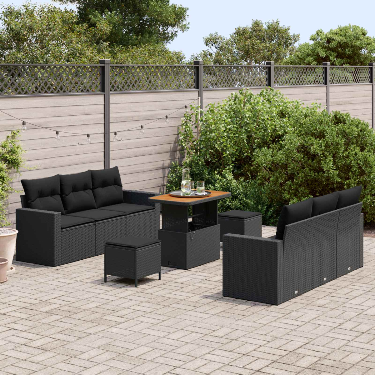 Garten-Set mit schwarzen Korbmöbeln, Sofas, Hockern und Tisch auf Steinplatten, mit Zaun und Grün.