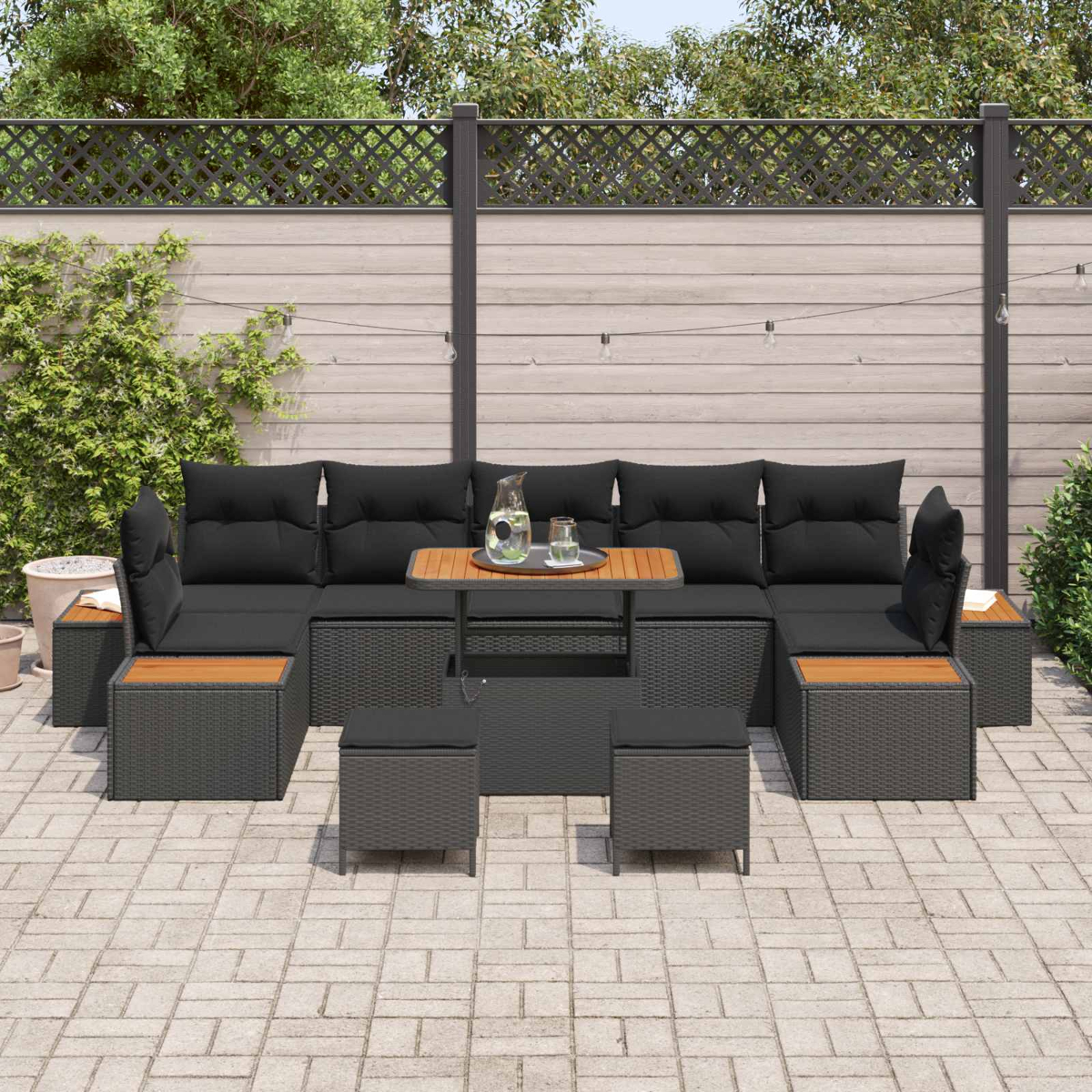 Set da patio esterno con cuscini neri, un tavolo in legno e piccoli pouf. Su un patio in mattoni con una recinzione sullo sfondo.