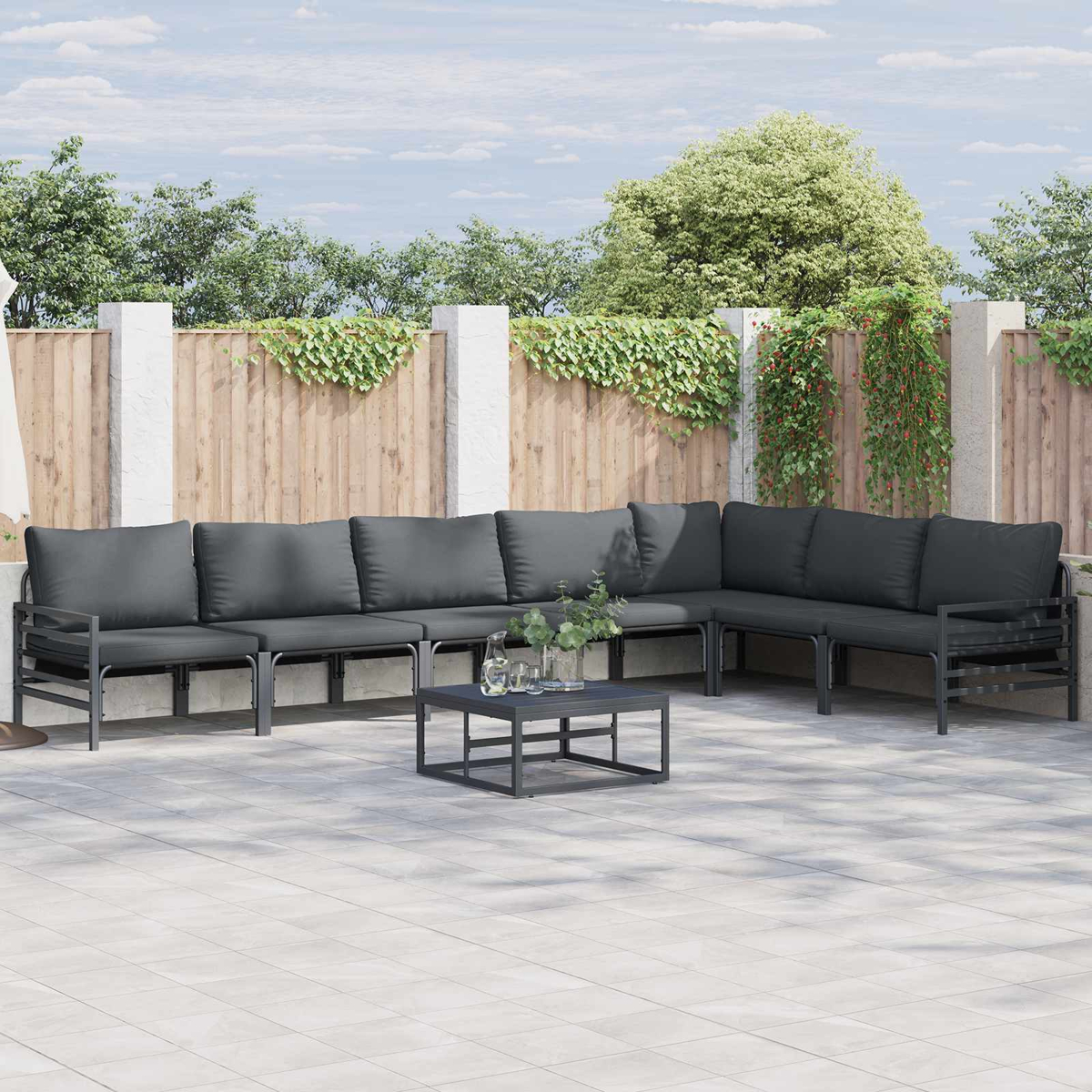 Gartensektionssofa mit dunkelgrauen Kissen, Couchtisch und Pflanzen auf einer gepflasterten Terrasse, Holzzaun im Hintergrund.