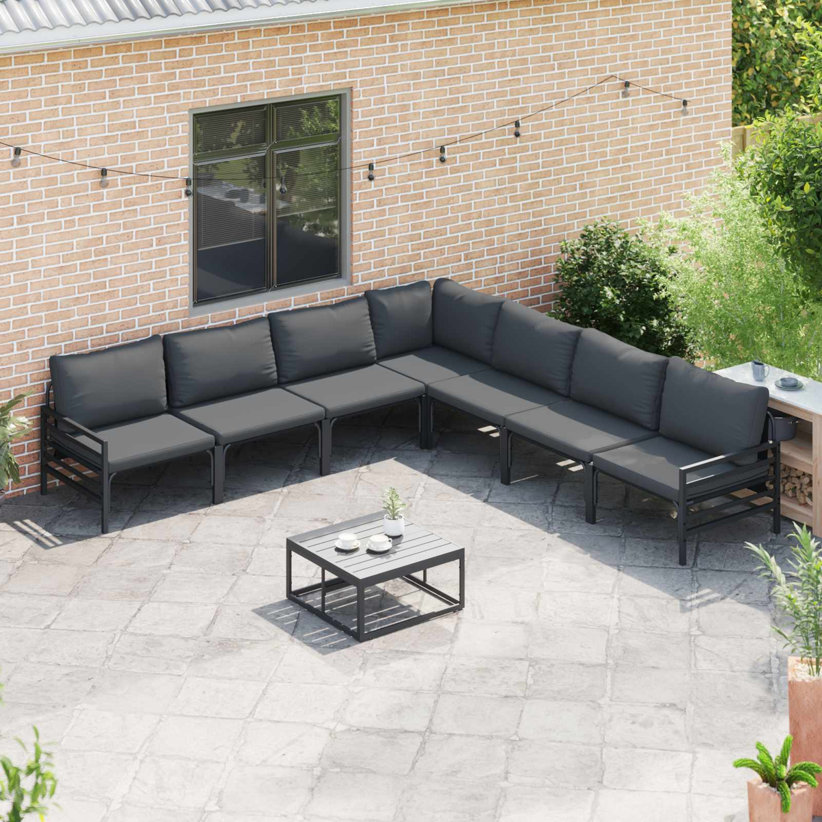 Outdoor-Terrassenset mit dunkelgrauen Kissen, kleinem Tisch, Backsteinmauer und grünen Pflanzen. Lichterketten sind oben gespannt.