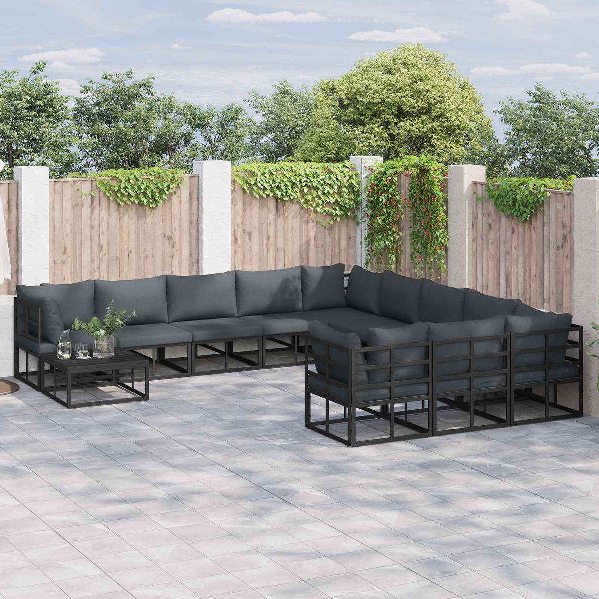 Outdoor-Ecksofa mit dunkelgrauen Kissen. Schwarzer Metallrahmen. Holzzaun im Hintergrund mit grünen Ranken und Bäumen.