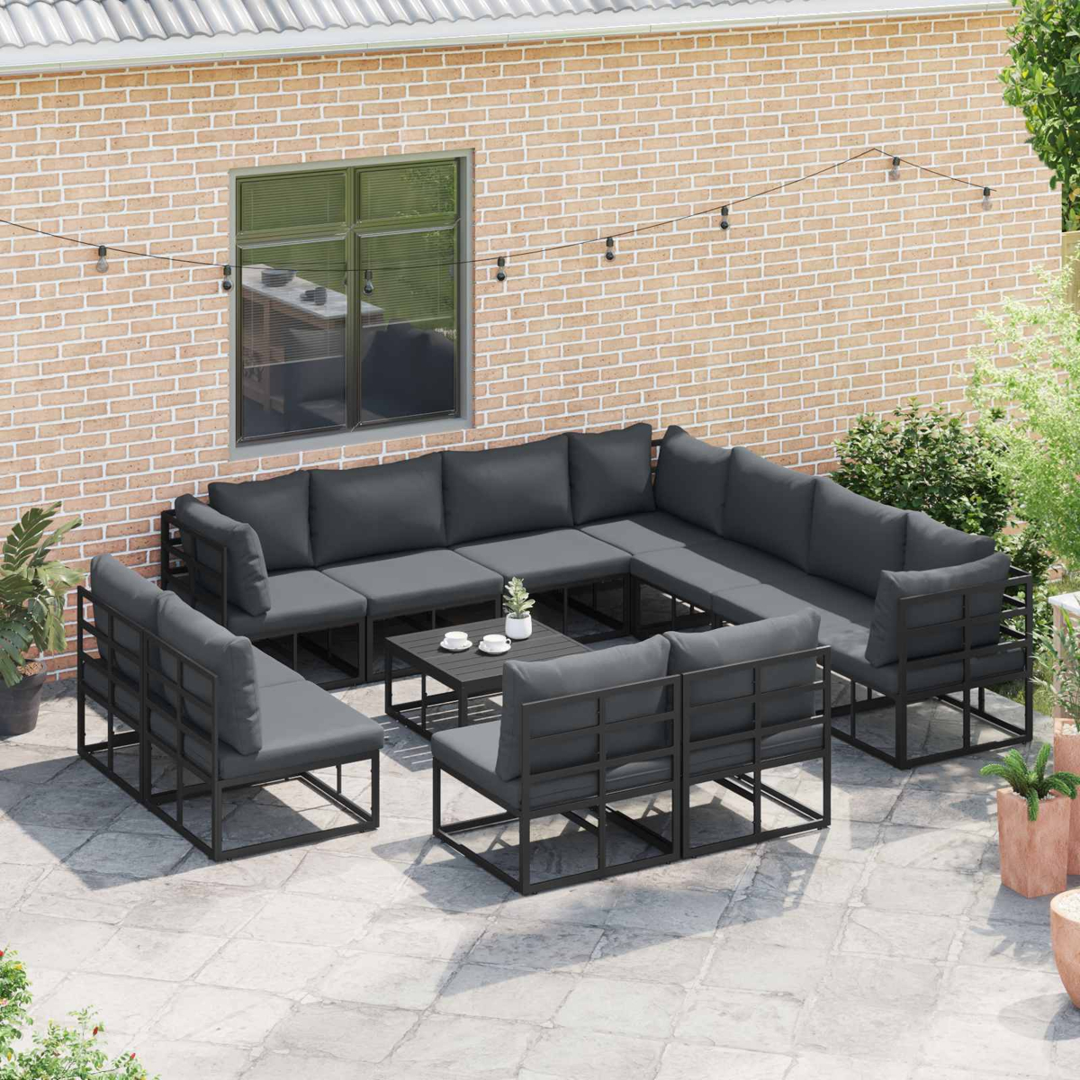 Gartensektionssofa mit grauen Kissen, einem schwarzen Couchtisch und Backsteinmauer im Hintergrund. Lichterketten und Pflanzen sind sichtbar.