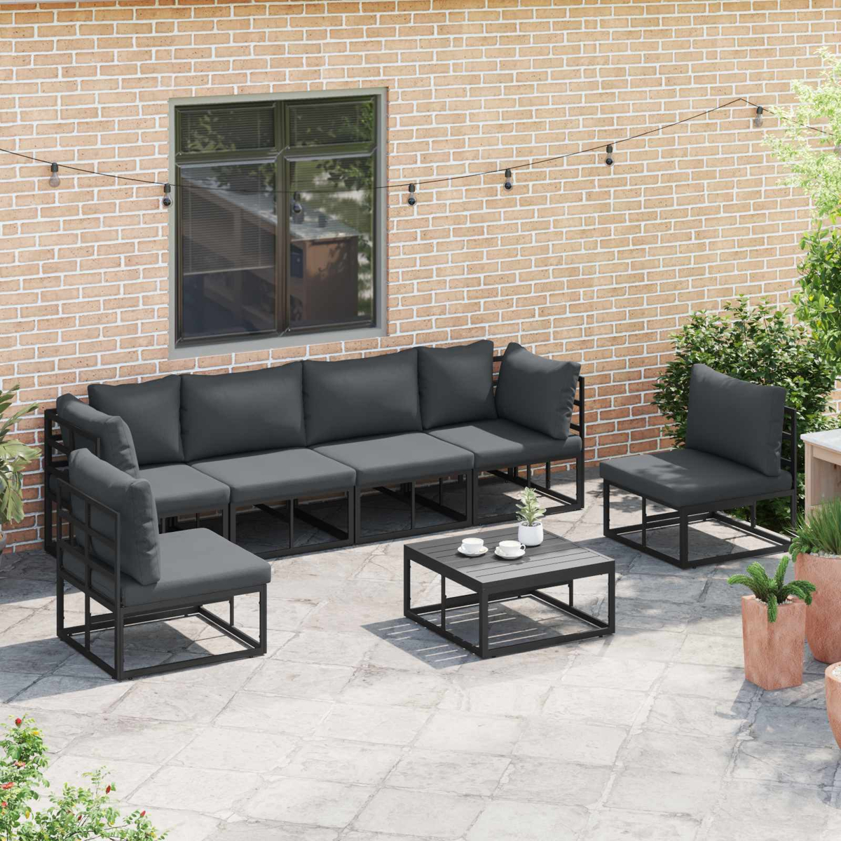 Terrassenset mit dunkelgrauen Kissen. Enthält ein Sofa, zwei Stühle und einen Couchtisch. Hintergrund ist eine Ziegelmauer, Fenster sichtbar.