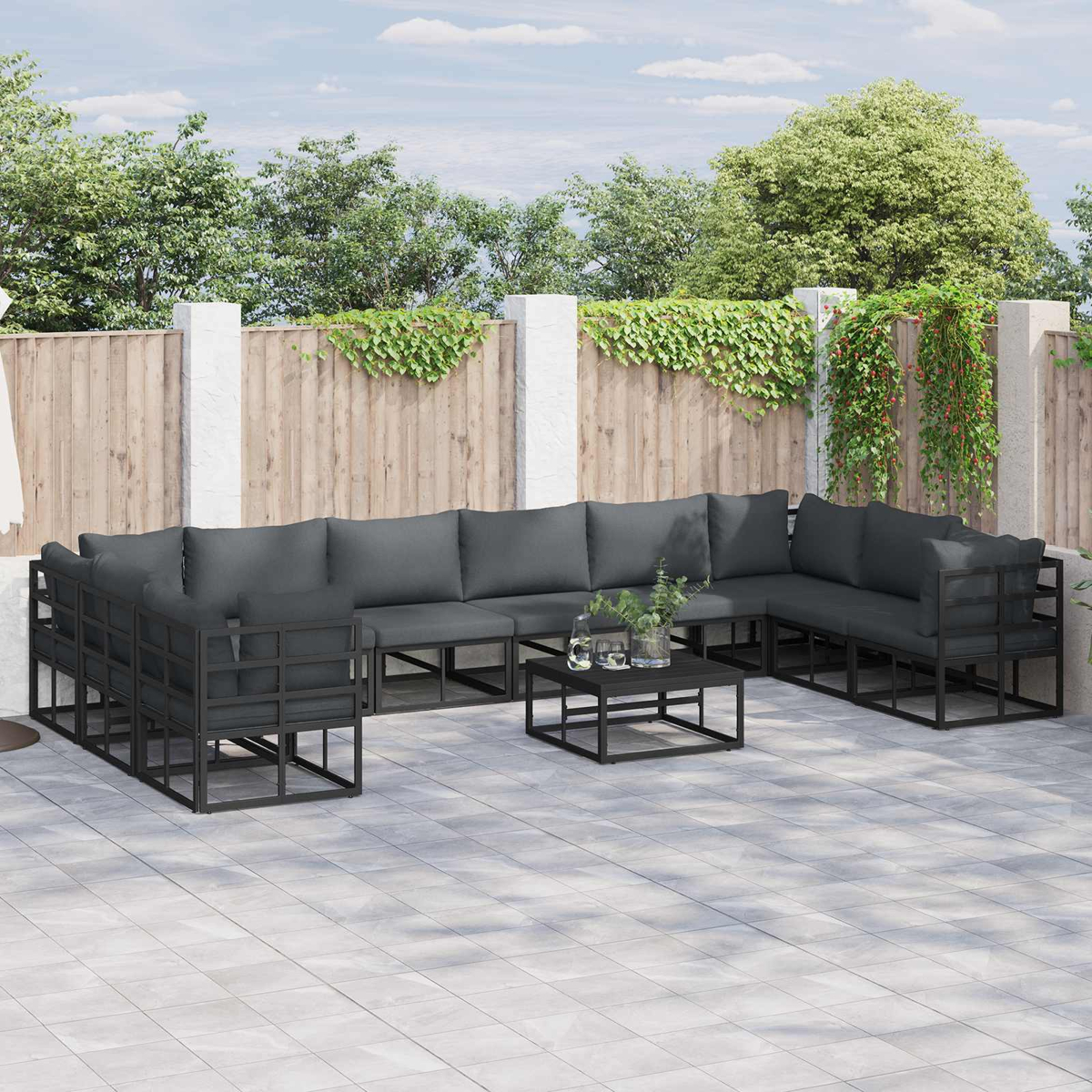 Terrassen-Set mit dunkelgrauen Kissen, schwarzem Metallrahmen und kleinem Tisch. Holzzaun und Grün im Hintergrund.