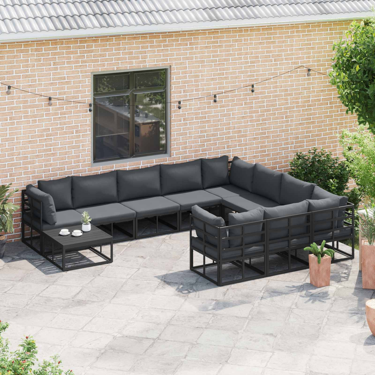 Outdoor-Ecksofa mit dunkelgrauen Kissen. Inklusive kleinem Tisch mit Tassen, Topfpflanzen und Backsteinmauer im Hintergrund.