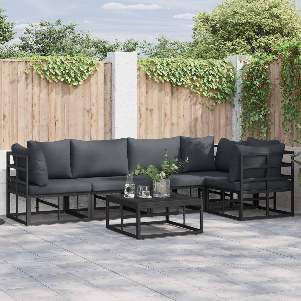 Gartenmöbel-Set mit grauen Kissen auf einer gefliesten Terrasse. Ein Holzzaun mit Kletterpflanzen bildet den Hintergrund.
