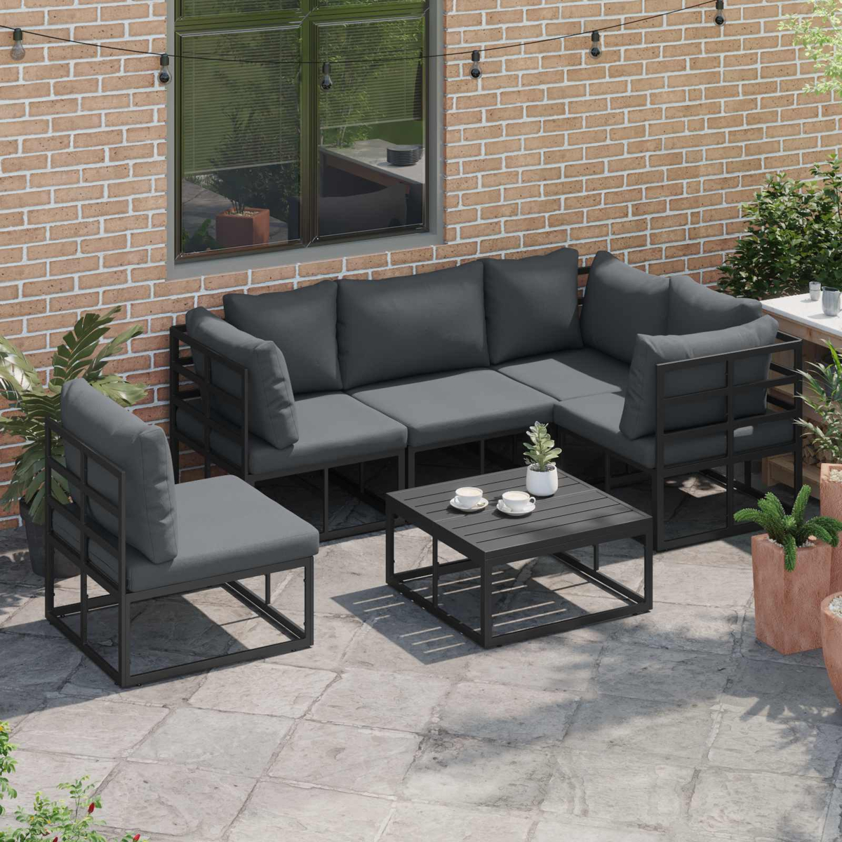 Garten-Set. Graue Kissen auf schwarzen Metallrahmen. Quadratischer Tisch mit Tassen und einer Pflanze. Ziegelwand im Hintergrund.