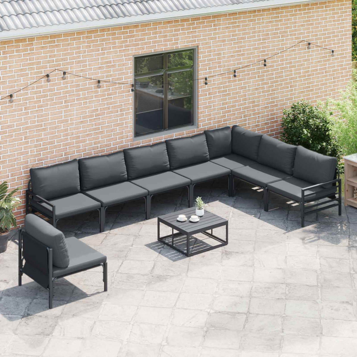 Set da patio esterno con divano componibile, tavolino e poltrona singola. Sfondo con muro di mattoni, luci a corda e una finestra.