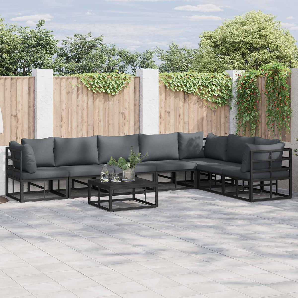 Outdoor-Ecksofa mit grauen Kissen und schwarzem Rahmen. Ein schwarzer Tisch mit Gläsern und Pflanzen davor. Holzzaun im Hintergrund.