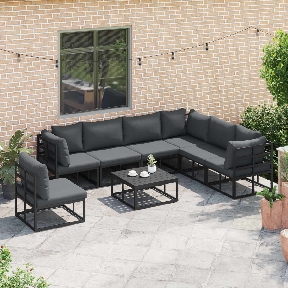 Terrassengarnitur mit Ecksofa, Stuhl und Couchtisch. Graue Kissen, schwarze Rahmen, Backsteinmauer und Pflanzen im Hintergrund.