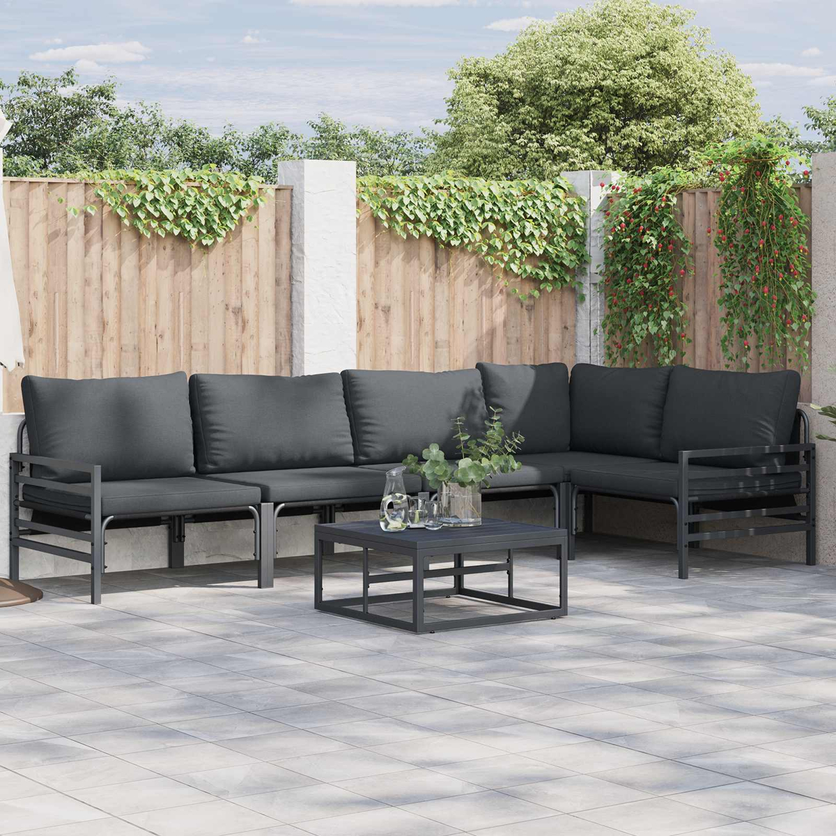 Garten-Sitzgruppe mit grauen Kissen und einem quadratischen Couchtisch auf einer gefliesten Terrasse. Holzzaun mit Kletterpflanzen im Hintergrund.