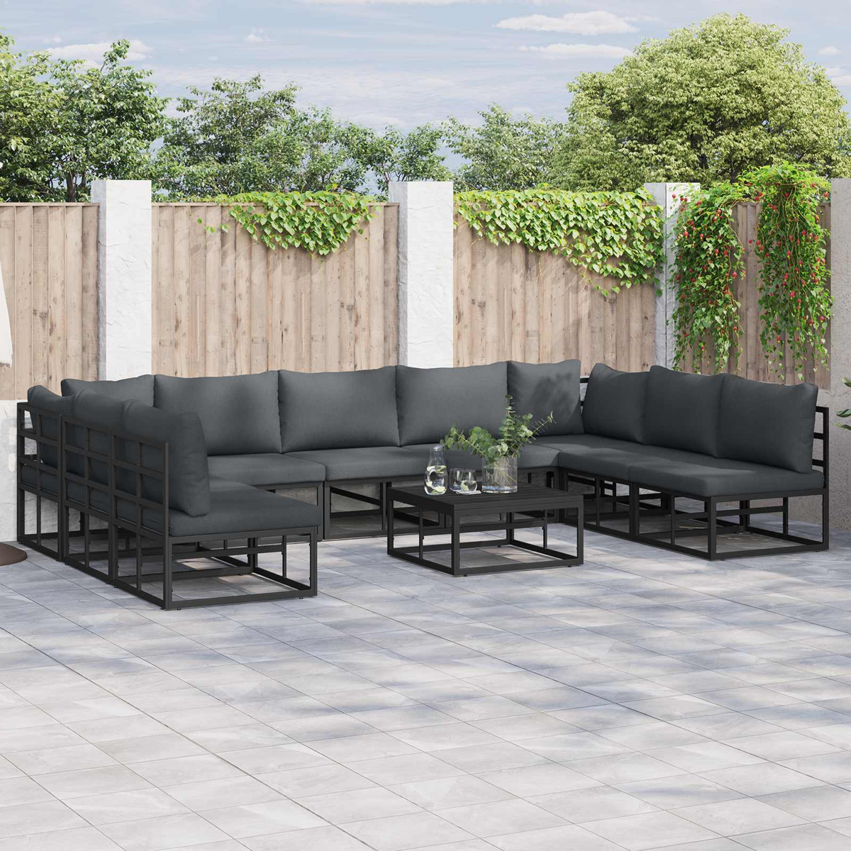 Outdoor-Ecksofa mit grauen Kissen auf einer gefliesten Terrasse. Ein kleiner Tisch hält Pflanzen und Getränke. Zaun und Grün im Hintergrund.