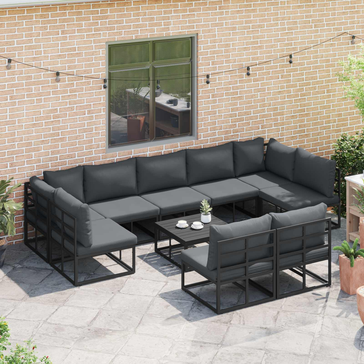 Terrassen-Set mit dunkelgrauen Kissen. Enthält ein Ecksofa, zwei Stühle und einen Couchtisch. Hintergrund aus Backsteinmauer.