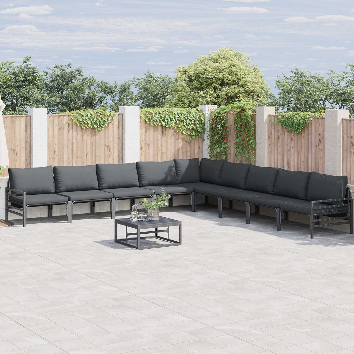Outdoor-Ecksofa mit grauen Kissen und einem kleinen Tisch auf einer Terrasse. Holzzaun und Grün im Hintergrund.