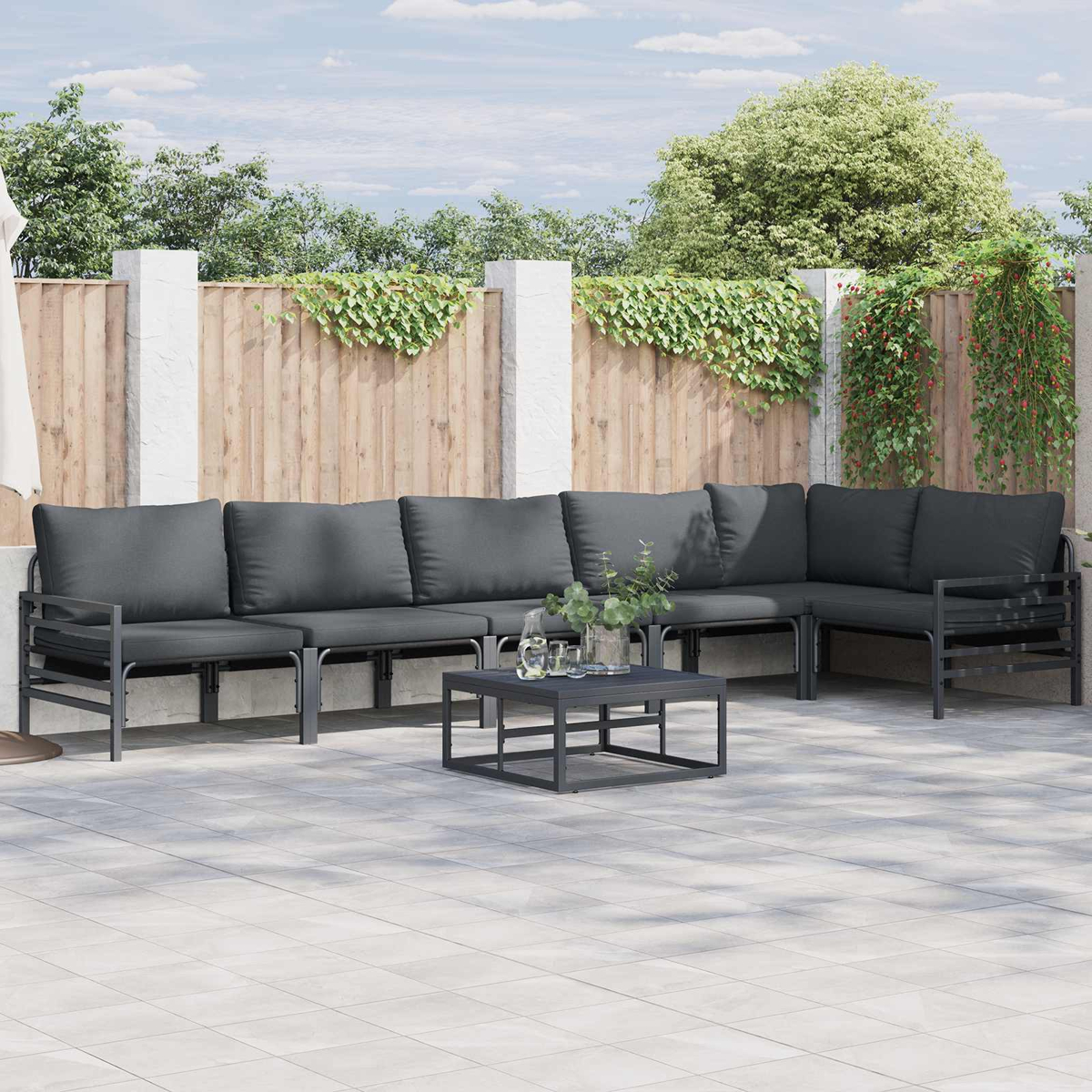 Garten-Sitzgruppe mit grauen Kissen und einem quadratischen Tisch. Hintergrund: Holzzaun und Grün.