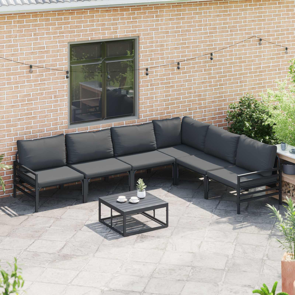 Outdoor-Ecksofa mit grauen Kissen und schwarzem Rahmen, kleiner Tisch mit Tassen, Hintergrund aus Ziegelmauer.