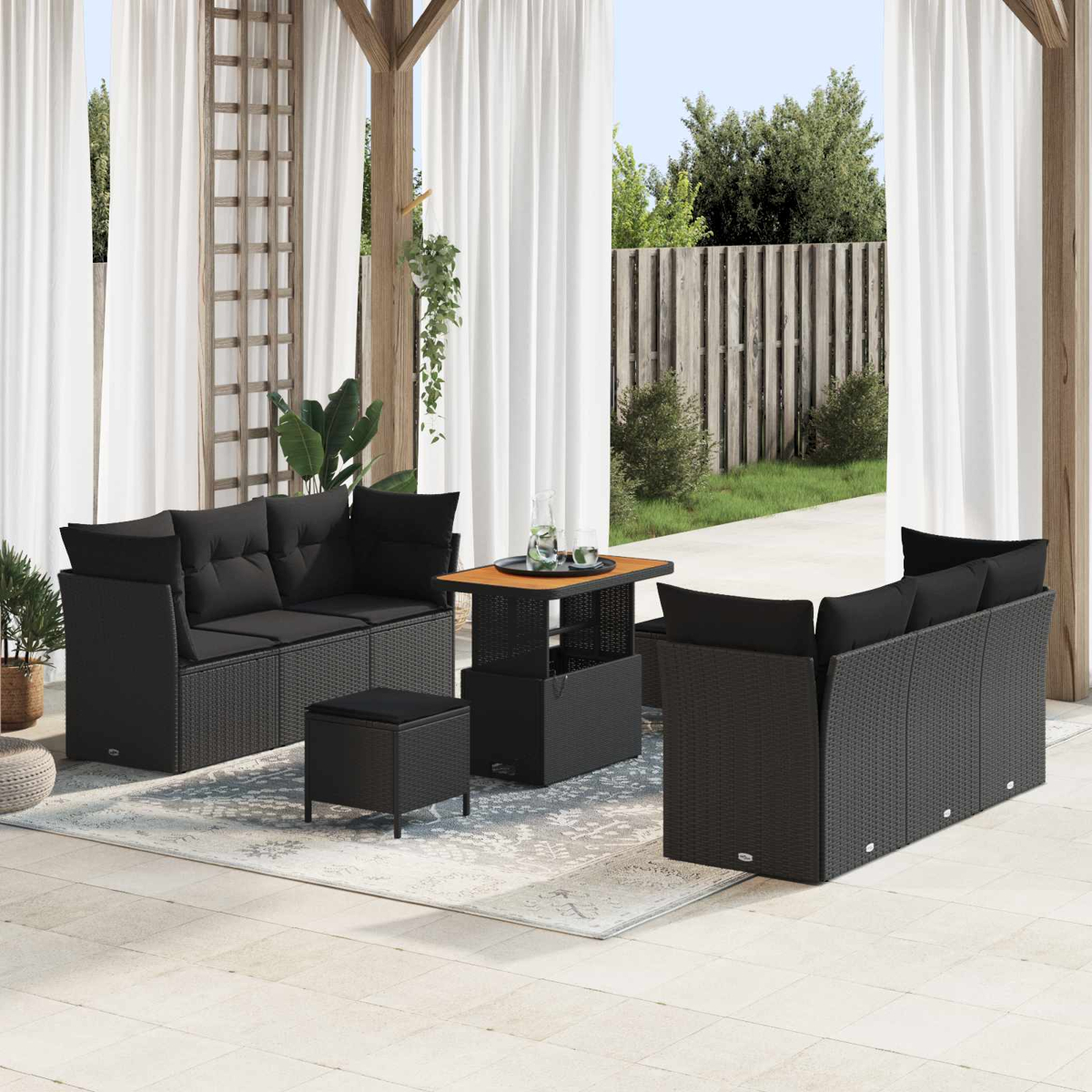 Terrassengarnitur mit schwarzen Korbmöbeln, darunter Sofas, ein Hocker und ein Tisch, auf einem gemusterten Teppich unter weißen Vorhängen.