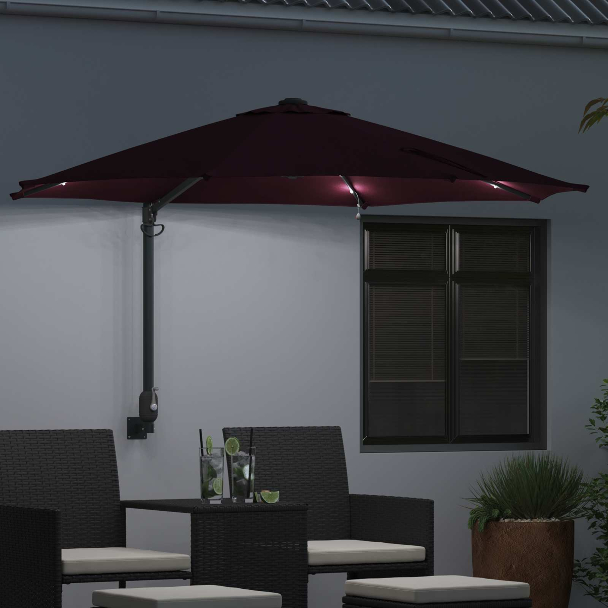 Un ombrellone esterno rosso scuro con luci. Sotto, un tavolo con bevande e sedie sul patio, con una finestra e piante.