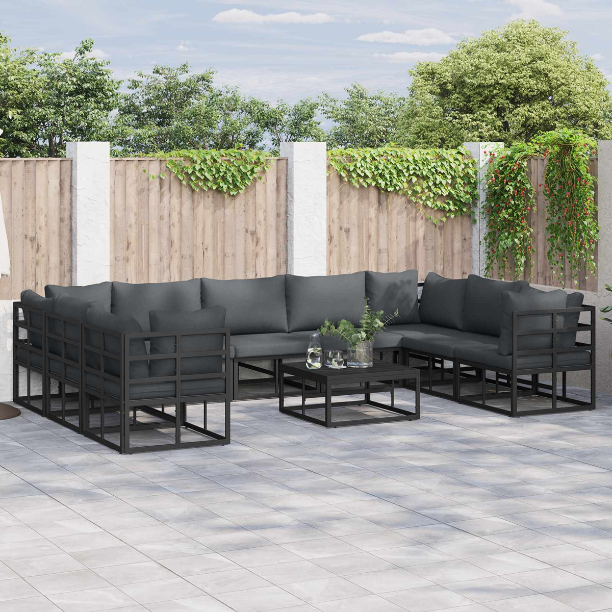 Set da patio esterno con cuscini grigio scuro, telai neri e un tavolino. Una recinzione in legno sullo sfondo.