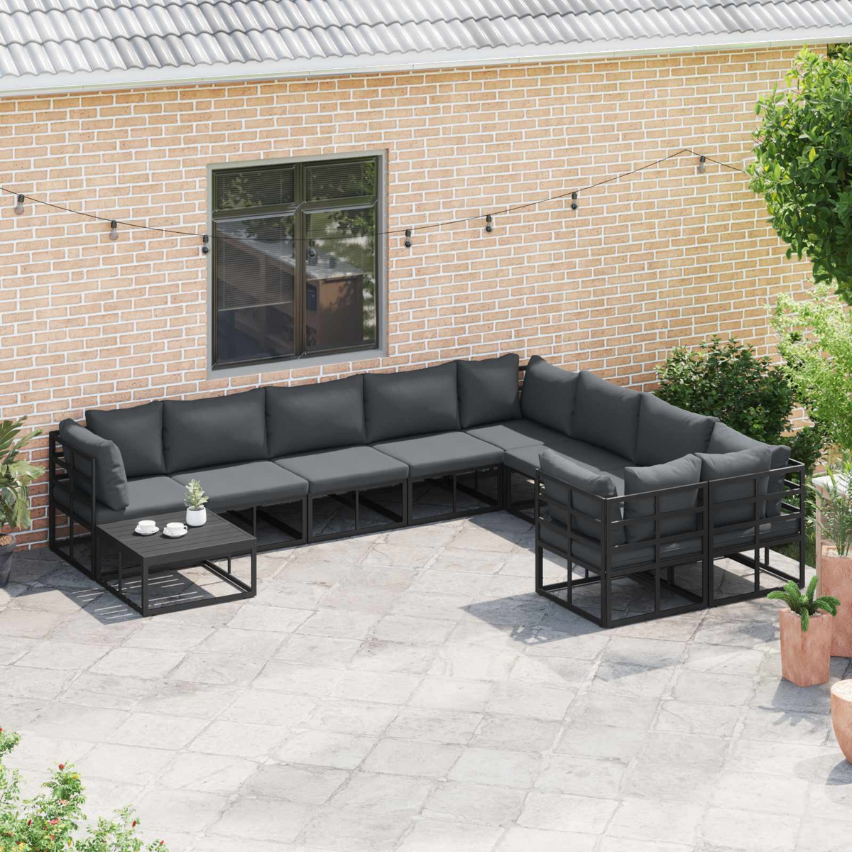 Set da patio all'aperto con cuscini grigio scuro. Include un divano componibile, un tavolino e un muro di mattoni sullo sfondo.