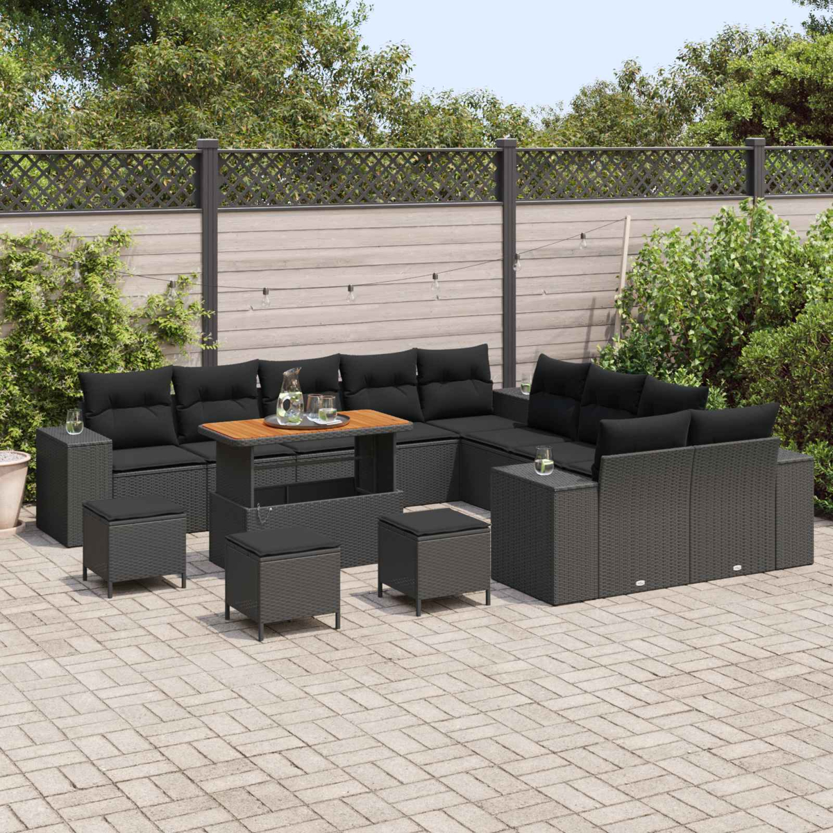 Garten-Terrassenset mit schwarzen Kissen und grauem Korbgeflecht. Enthält einen Tisch, Hocker und Sofas. Hintergrund: Zaun und Grün.