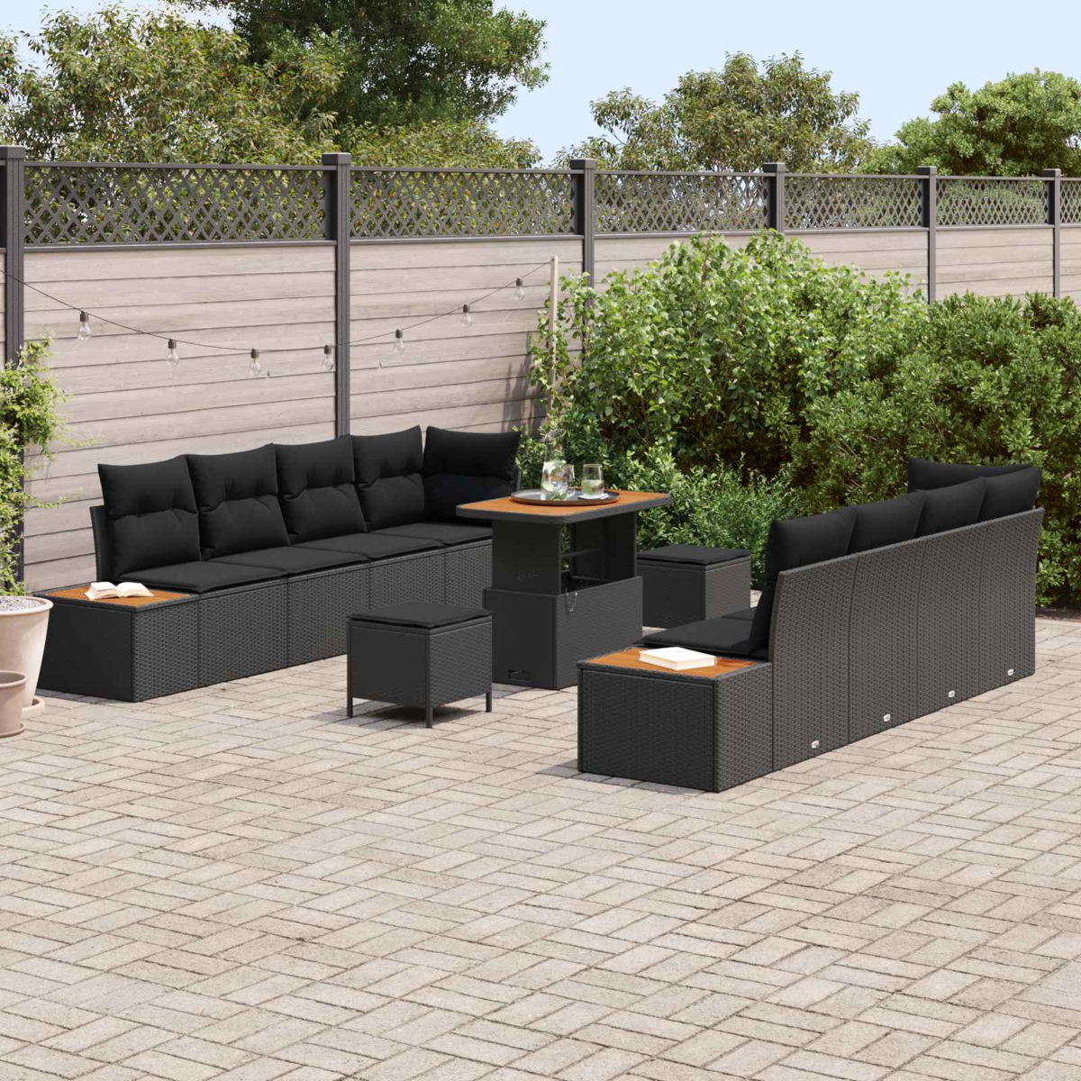 Terrassengarnitur mit schwarzer Korbmöbeln, Tisch mit Holzplatte und passenden Hockern auf einer Steinpflasterterrasse mit Zaun.
