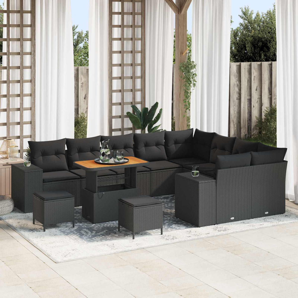 Sofá seccional para exteriores con cojines negros y una mesa de madera. Cortinas blancas y un enrejado de madera en el fondo.