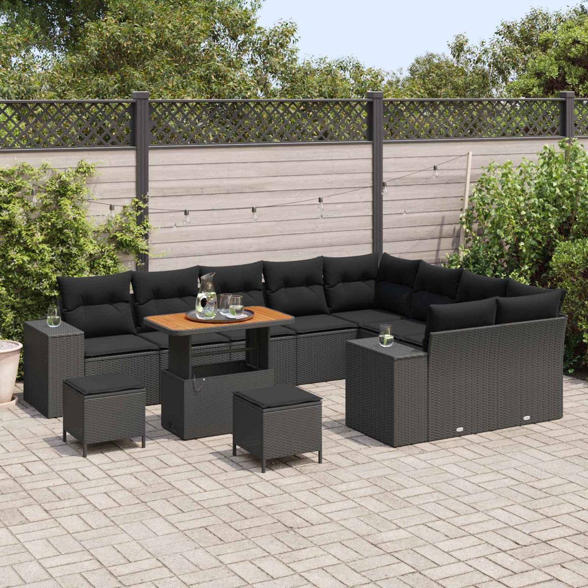 Sofá seccional exterior con cojines negros, mesa de madera y otomanas en un patio. Valla de madera y vegetación al fondo.