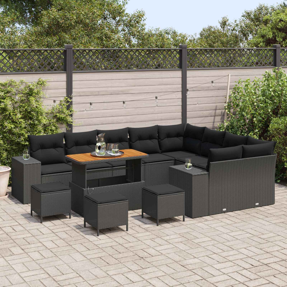 Garten-Terrassenset mit schwarzen Kissen, Holztisch und grau gewebten Möbeln. Auf einer gepflasterten Terrasse mit Holzzaun im Hintergrund.