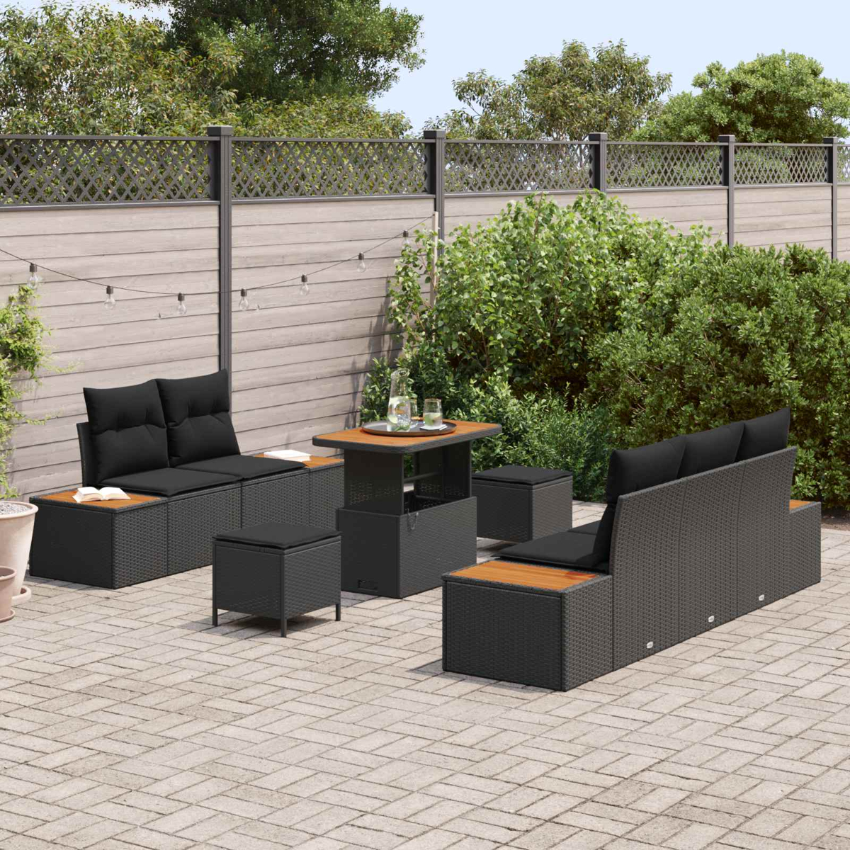 Garten-Terrassenset mit schwarzen Korbmöbeln, Holztischplatten und schwarzen Kissen auf einer Steinpflasterterrasse, mit Grün im Hintergrund.