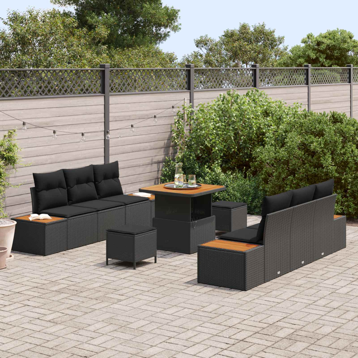 Terrassen-Set mit schwarzen Kissen und Holztisch. Sofa, Stühle und Hocker auf Steinterrasse mit Grün.