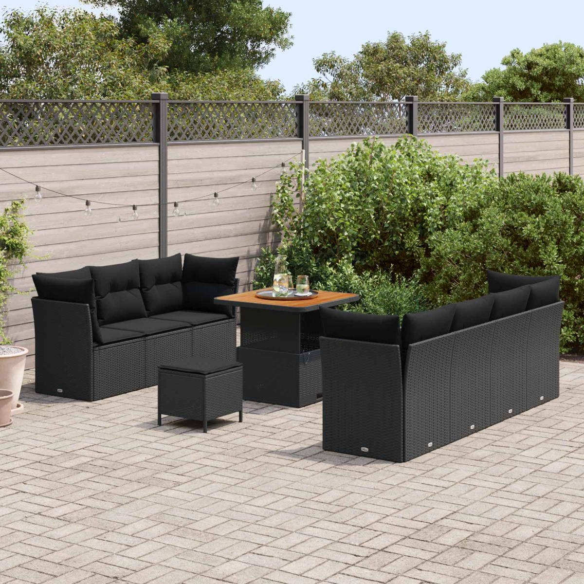 Set da patio esterno con divani in vimini neri, un tavolo e un piccolo pouf. La scena comprende una recinzione e vegetazione.