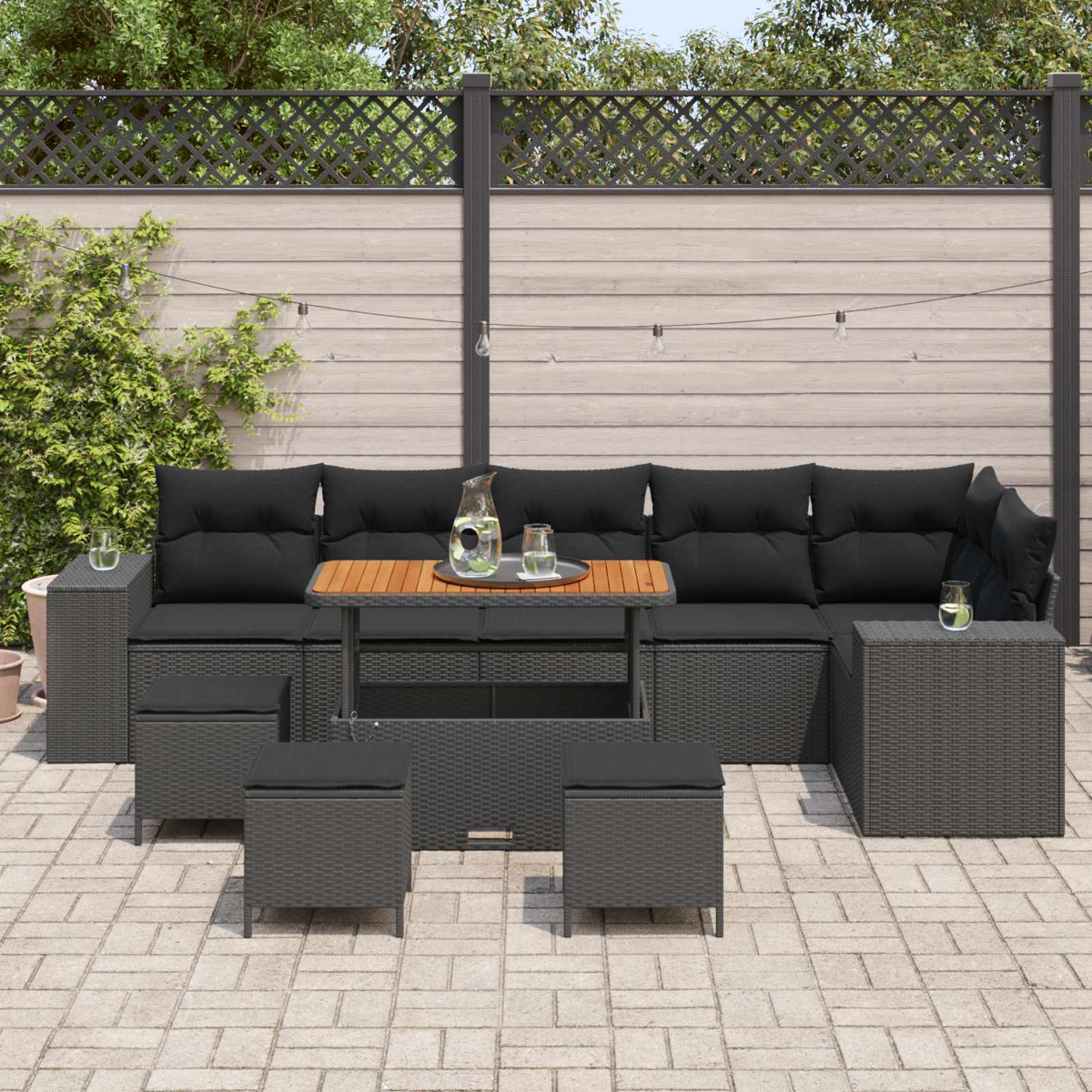 Terrassengarnitur mit schwarzen Kissen und Holztisch. Enthält Sofa, Hocker und Beistelltisch. Hintergrund: Zaun und Pflanzen.