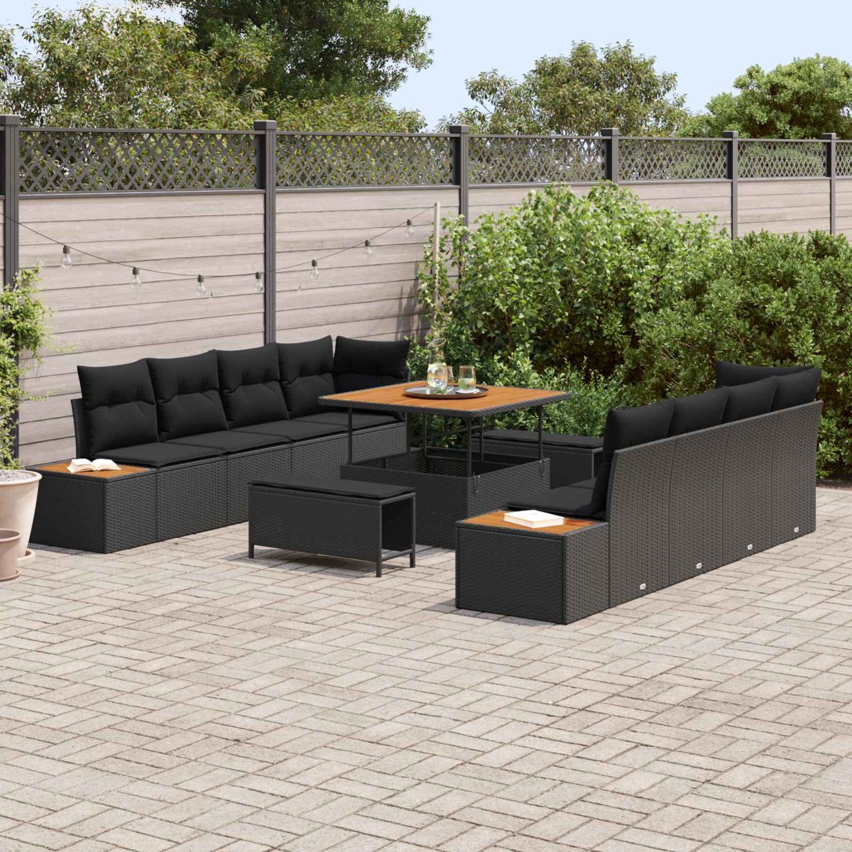 Terrassen-Set mit schwarzen Korbmöbeln, Holztisch und Kissen. Auf einer Steinterrasse mit Zaun und Grün.