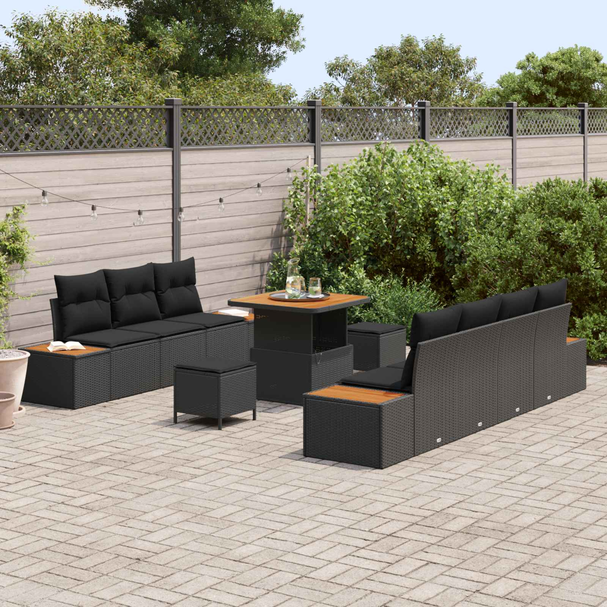 Garten-Set mit schwarzem Korbmöbel, Holztisch und Kissen. Auf einer Backstein-Terrasse mit Zaun und Grün im Hintergrund.