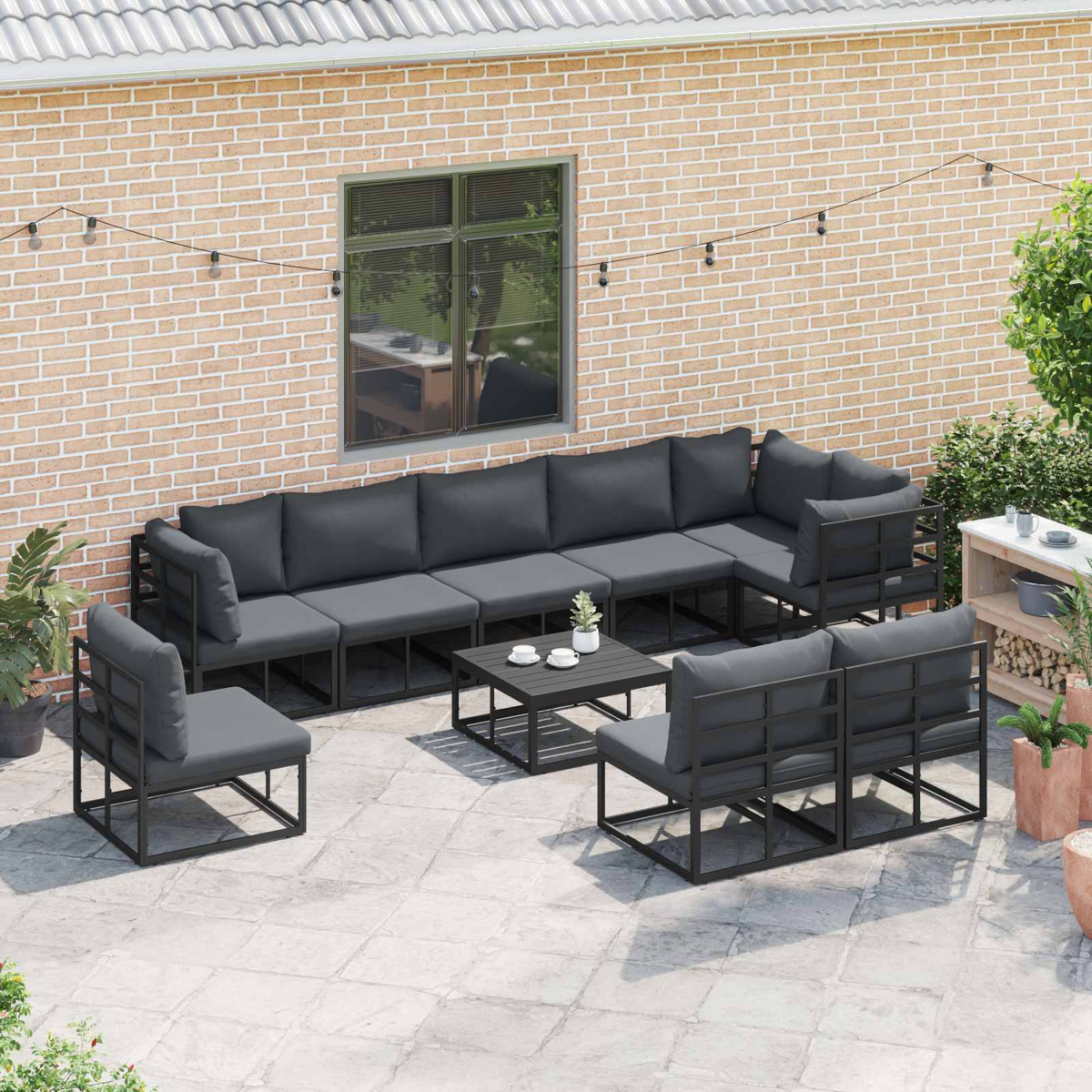 Set da patio esterno con cuscini grigio scuro. Include un divano componibile, sedie e un tavolino. Contro un muro di mattoni.