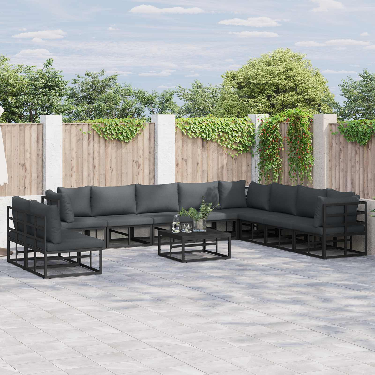 Outdoor-Ecksofa mit dunkelgrauen Kissen. Ein schwarzer quadratischer Tisch. Holzzaun mit grünen Pflanzen im Hintergrund.