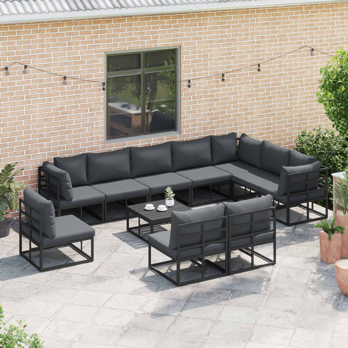 Garten-Terrassenset mit dunkelgrauen Kissen, angeordnet auf einem gepflasterten Bereich, vor einer Backsteinmauer. Enthält einen Tisch und Lichterketten.
