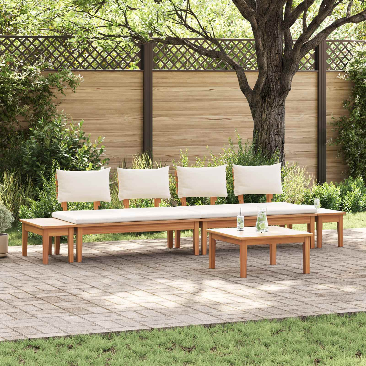 Outdoor-Sitzbereich mit Holzbänken, weißen Kissen und einem Tisch. Ein Baum und ein Zaun sind im Hintergrund. Getränke stehen auf dem Tisch.
