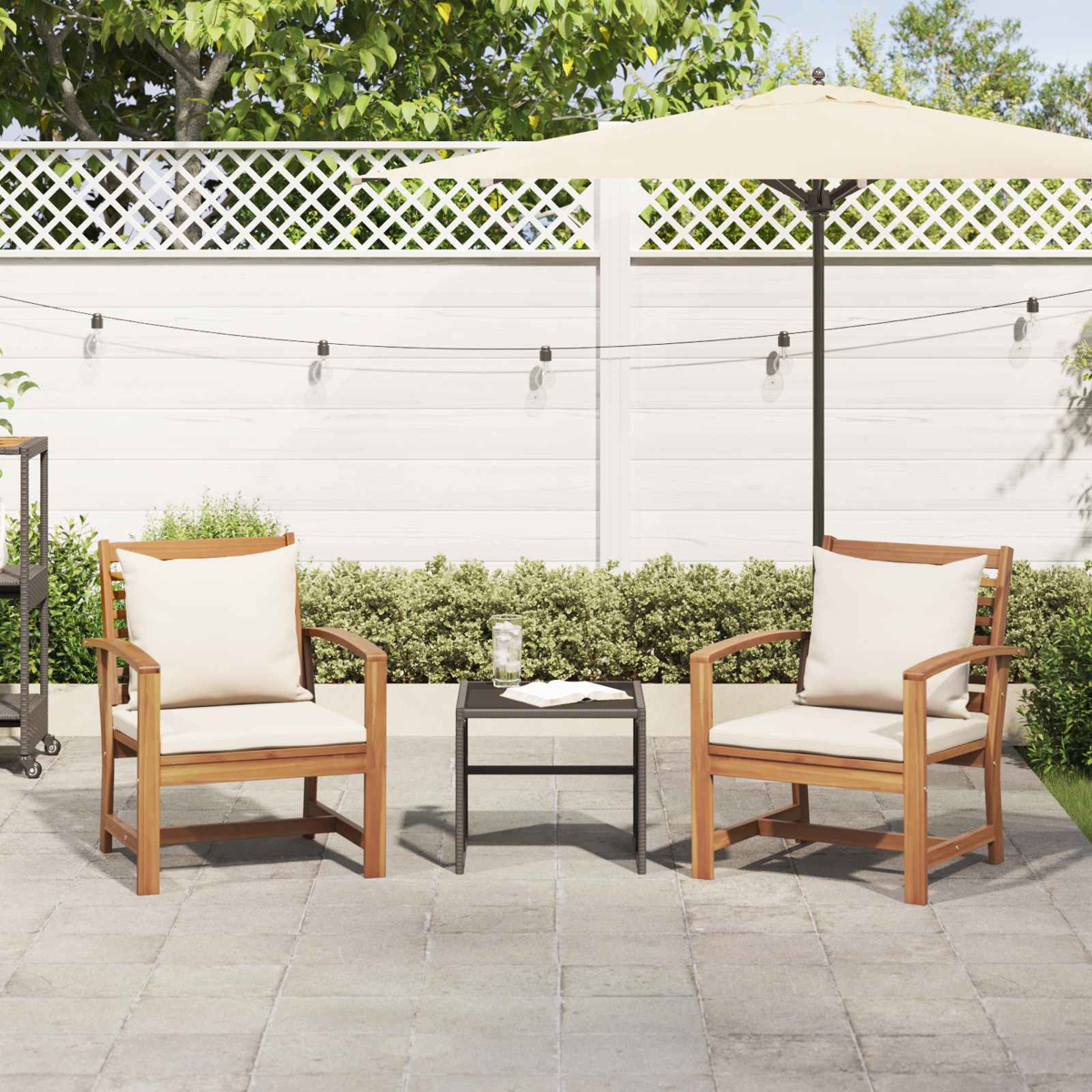 Dos sillas de madera con cojines blancos y una mesa pequeña en un patio. Hay una sombrilla y una valla blanca al fondo.