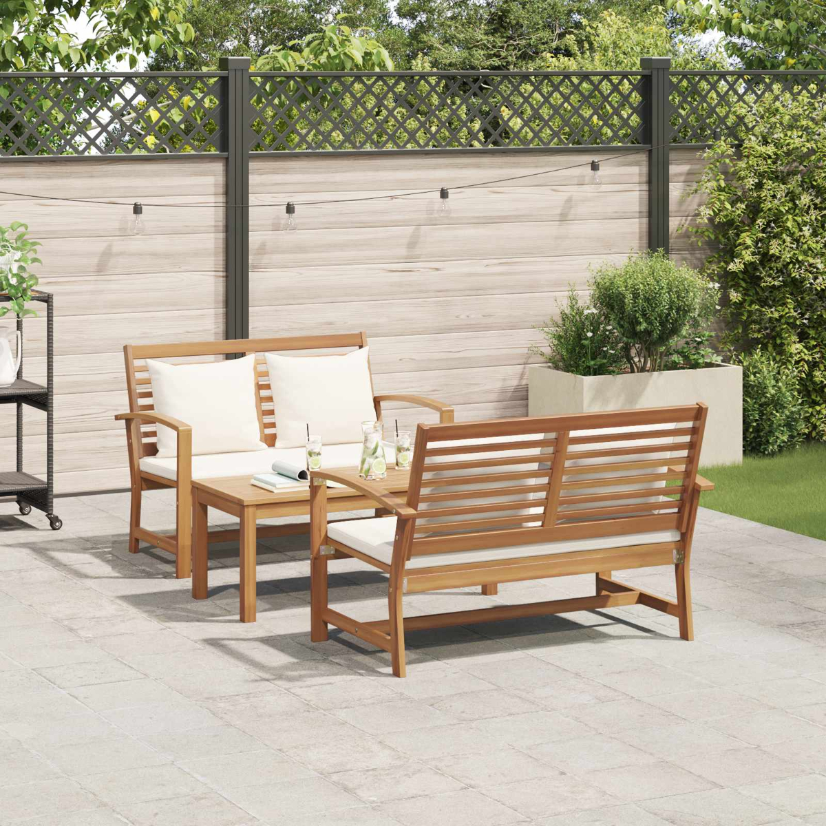 Juego de patio al aire libre con muebles de madera, cojines blancos y una mesita. Una valla y plantas al fondo.