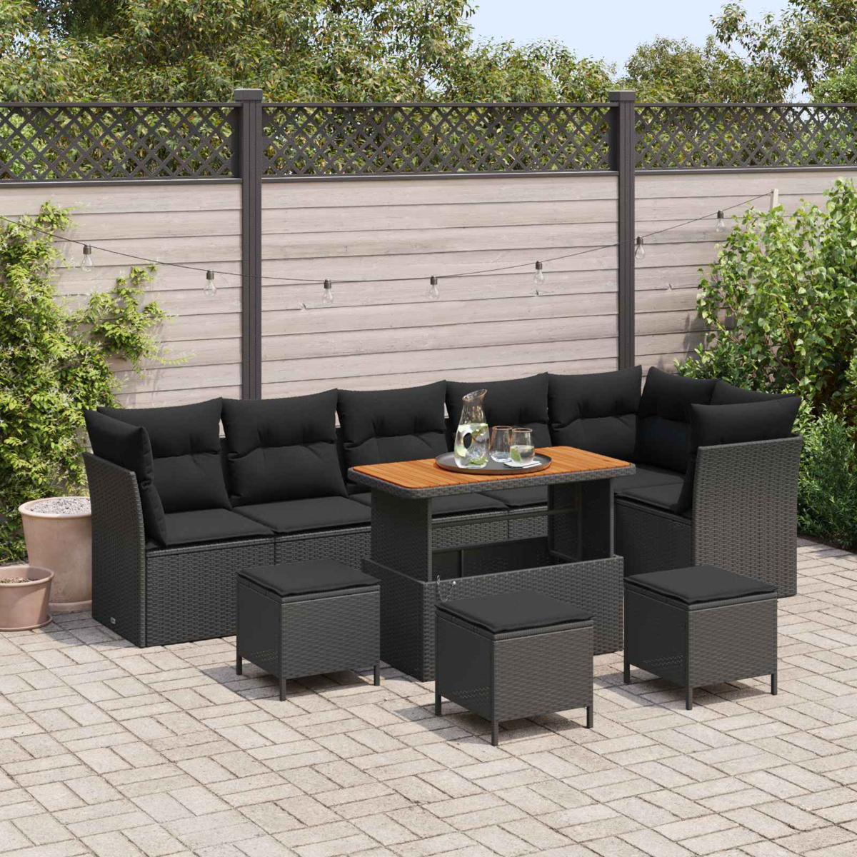 Terrassengarnitur im Freien mit schwarzen Kissen, einem Holztisch und Hockern. Der Hintergrund zeigt einen Zaun und Grün.